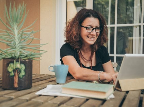 Middle aged female studying at home with books, newspaper and digital tablet pad. Woman reading a book and watching video online on new tech device. Education, modern lifestyle and leisure concept.