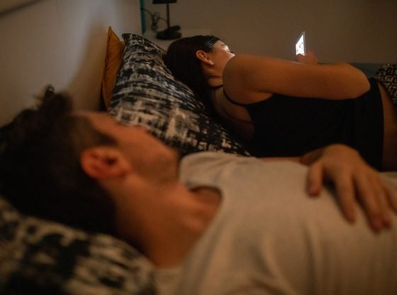 Young woman using phone in bed while her husband is asleep